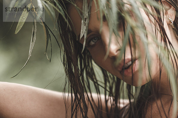 Nahaufnahme einer jungen Frau mit nassen Haaren,  die durch die Blätter schaut.