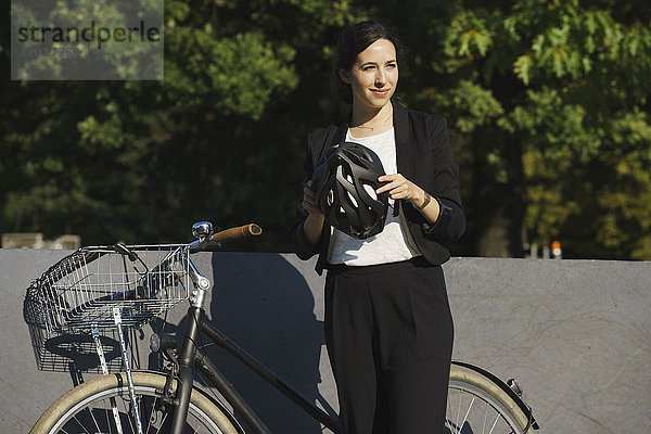 Lächelnde Geschäftsfrau,  die an einem sonnigen Tag den Helm am Fahrrad gegen die Pflanzen hält.