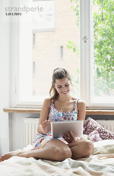 Schöne Frau mit digitalem Tablett,  während sie zu Hause auf dem Bett gegen das Fenster sitzt.