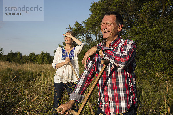 Lächelndes reifes Paar mit Ausrüstung,  das an einem sonnigen Tag auf dem Bauernhof steht.