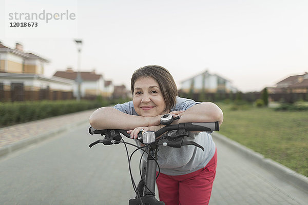 Porträt einer älteren Frau,  die sich auf der Straße über den Fahrradgriff lehnt.