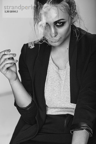Porträt einer jungen Frau,  die im Sitzen vor grauem Hintergrund eine Zigarette raucht.