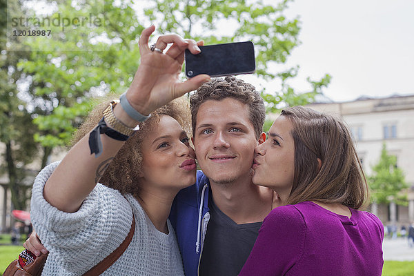 Frau nimmt Selfie mit einer Freundin,  während sie einen jungen Mann küsst.