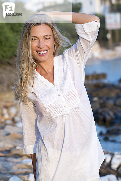 Porträt einer lächelnden jungen blonden Frau im weißen Hemd auf einem Felsen im Meer.