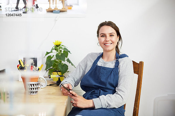 Porträt einer lächelnden jungen Ingenieurin mit Smartphone am Tisch in der Werkstatt