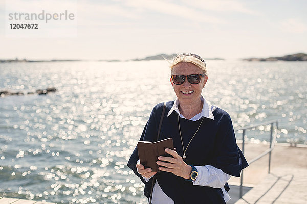 Porträt einer lächelnden Seniorin mit Sonnenbrille,  die ihr Handy gegen den See hält.