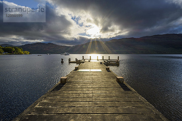 Holzsteg am See mit dramatischen Wolken bei Sonnenaufgang am Loch Lomond in Schottland,  Vereinigtes Königreich