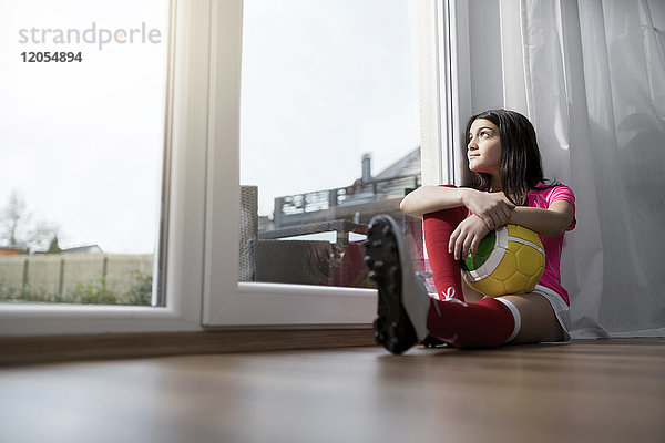 Mädchen im Fußball-Outfit sitzend auf dem Boden im Wohnzimmer mit Blick aufs Fenster
