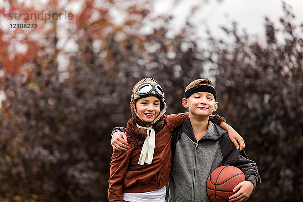 Porträt eines Mädchens und Zwillingsbruders in Basketballer- und Pilotenkostümen zu Halloween im Park