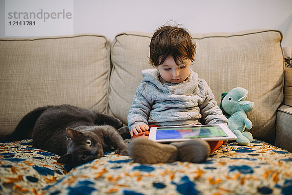 Kleines Mädchen sitzt auf der Couch und schaut sich Videos auf einem Tablett mit einer Katze an.