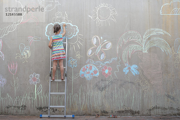 Mädchen steht auf einer Leiter und zeichnet bunte Bilder mit Kreide auf einer Betonwand.