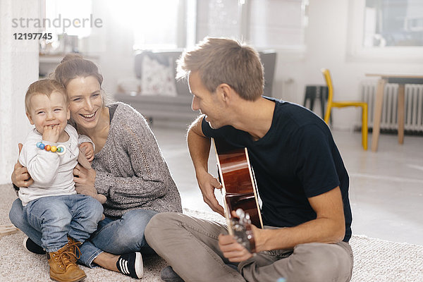 Glückliche Familie sitzt auf dem Boden und Vater spielt Gitarre.
