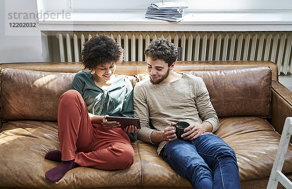 Junger Mann und junge Frau sitzend auf der Couch mit Tasse Kaffee und Tablette