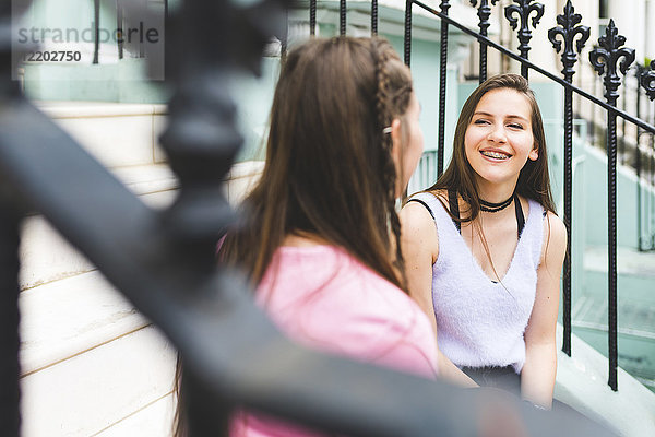 Zwei glückliche Teenager,  die auf einer Treppe in der Stadt sitzen und reden.