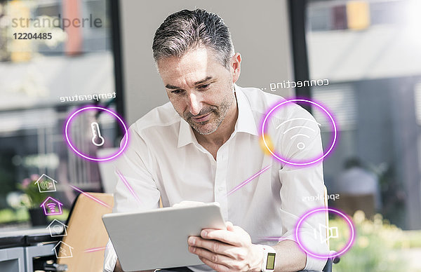 Ein Mann sitzt im Büro und benutzt ein digitales Tablett,  um sein intelligentes Zuhause zu steuern.