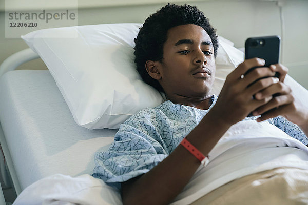 Schwarzer Junge liegt im Krankenhausbett und schreibt eine SMS mit seinem Handy