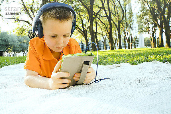 Ernster kaukasischer Junge,  der auf einer Decke im Park liegt und einem digitalen Tablet zuhört