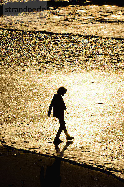 Silhouette eines gemischtrassigen Jungen,  der bei Sonnenuntergang am Strand spazieren geht