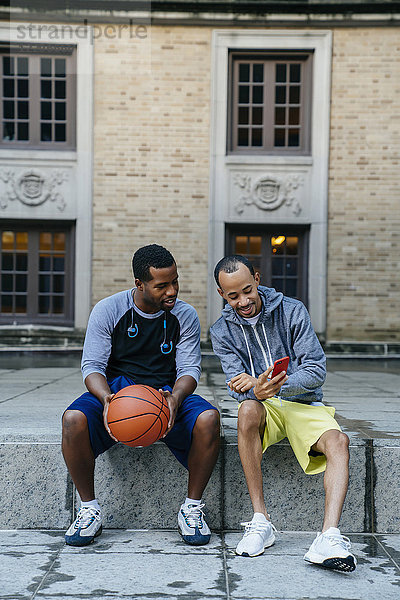 Schwarze Männer,  die mit einem Basketball auf dem Beton sitzen und mit dem Handy SMS schreiben