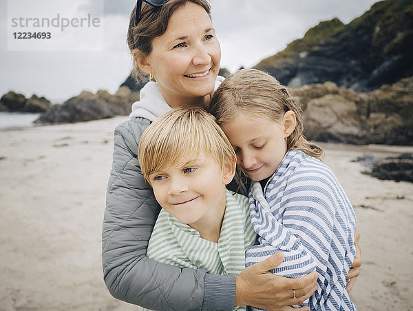 Lächelnde Frau umarmt Kinder,  die am Strand stehen.