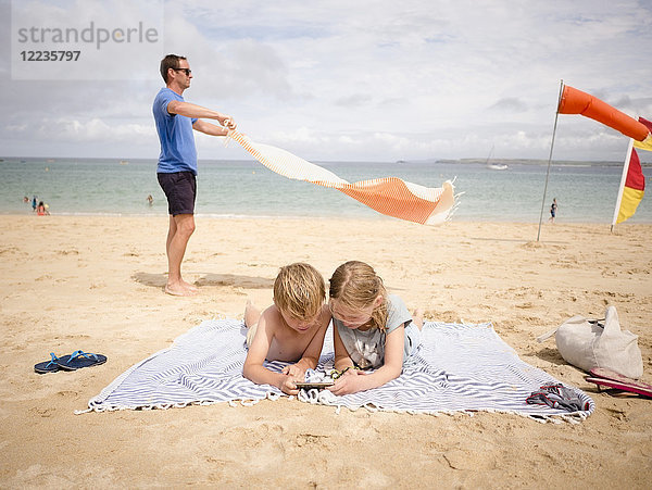 Mann hält Handtuch,  während Kinder sich am Strand ein Smartphone gegen den Himmel teilen.