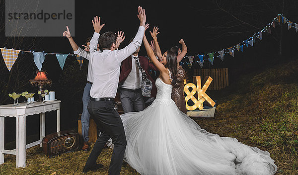Fröhliche Braut und Freunde mit erhobenen Armen tanzen und Spaß haben auf einer nächtlichen Feldparty.