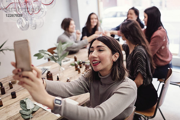Lächelnde junge Frau nimmt Selfie mit weiblichen Kollegen am Tisch in der Parfümwerkstatt.
