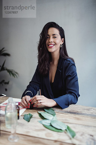 Porträt einer lächelnden,  erwachsenen Unternehmerin am Tisch gegen die Wand in der Parfumwerkstatt.
