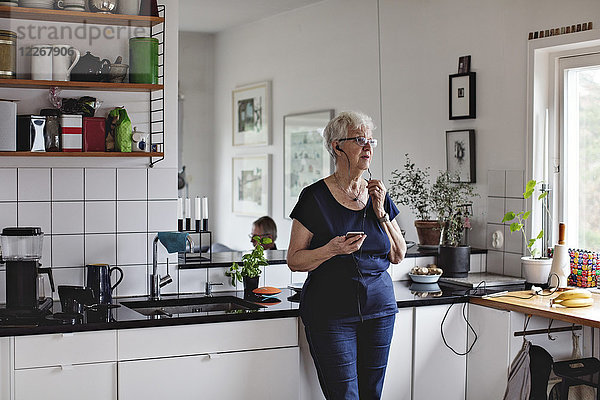 Seniorin im Ruhestand mit Smartphone beim Hören von In-Ear-Kopfhörern in der heimischen Küche