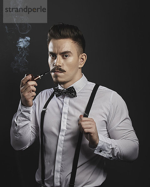 Porträt eines selbstbewussten jungen Mannes mit Hosenträgern beim Pfeifenrauchen über grauem Hintergrund
