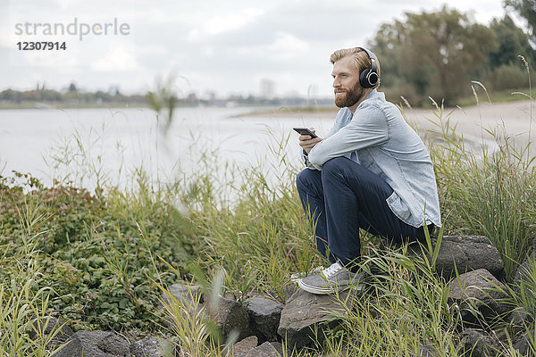 Deutschland,  Düsseldorf,  Mann hört Musik mit Kopfhörer und Smartphone in der Natur