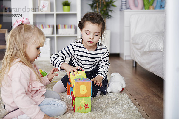 Zwei kleine Mädchen,  die zu Hause mit Spielzeugblöcken auf dem Teppich spielen.