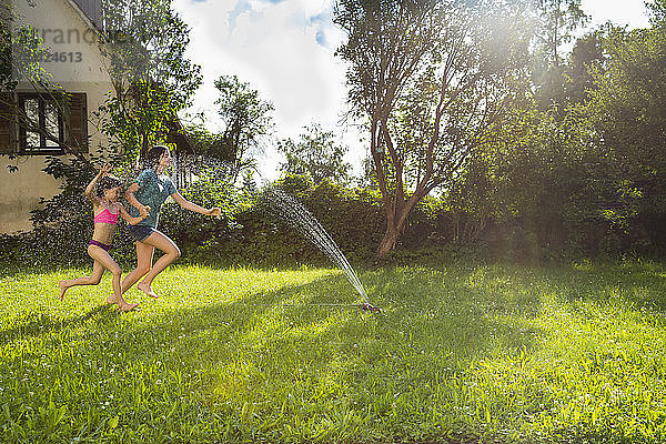 Girl and her little sister having fun with lawn sprinkler in the garden