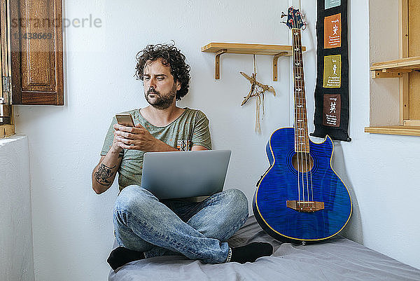 Man in his room with mobile phone,  laptop and bass guitar