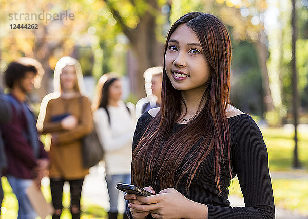 Porträt einer jungen internationalen Universitätsstudentin,  die ein Smartphone in der Hand hält,  mit ihren Freunden im Hintergrund,  Edmonton,  Alberta,  Kanada