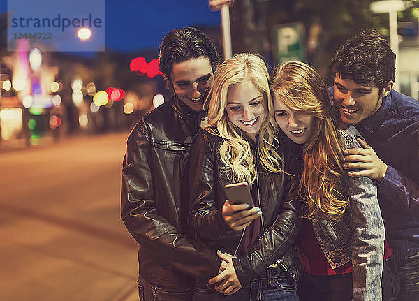 Eine Gruppe von vier Freunden kauert auf einem Bürgersteig zusammen und schaut auf ein Smartphone,  während das Leuchten des Bildschirms ihre Gesichter erhellt,  Edmonton,  Alberta,  Kanada