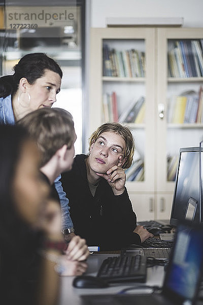 Confident female mature teacher discussing with male students while looking at computer monitor in classroom