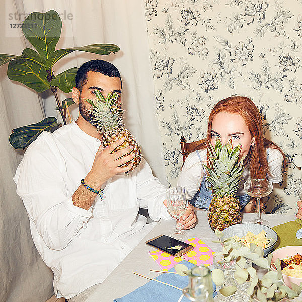 Portrait of playful young male and female friends with pineapples sitting at dining table during party in apartment