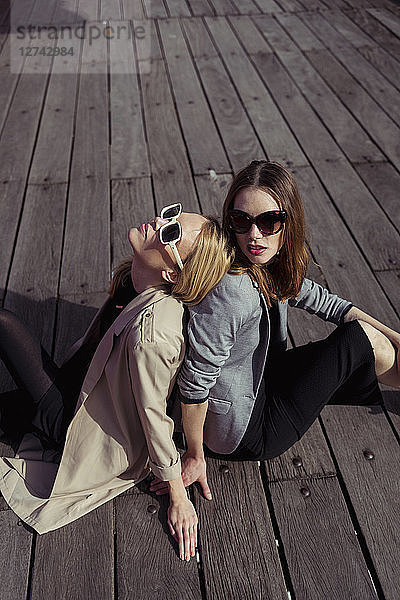 Portrait of two fashionable young women sitting on wooden floor wearing sunglasses