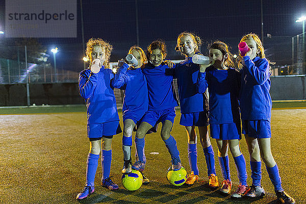 Porträt einer selbstbewussten Mädchenfußballmannschaft,  die nachts auf dem Spielfeld Wasser trinkt