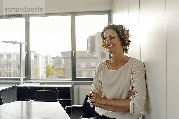 Businesswoman leaning against wall in office,  with arms crossed,  smiling