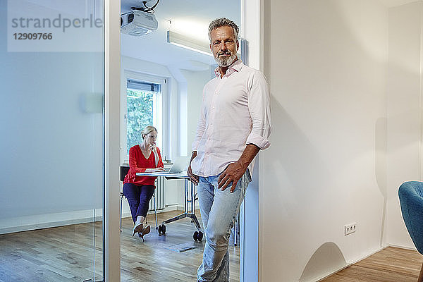 Portrait of mature man leaning against doorframe in office with colleague in background