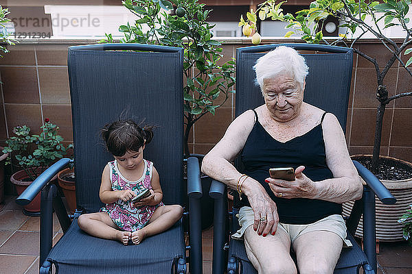 Grandmother and baby girl sitting side by side on the terrace using mobile phones