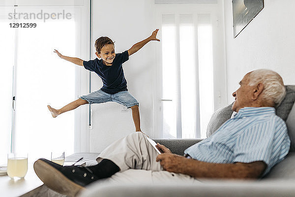 Grandfather sitting on the sofa with tablet watching his grandson jumping in the air