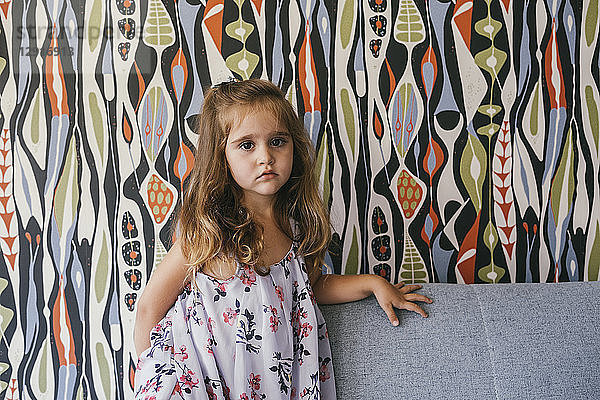 Portrait of serious little girl on couch in front of patterned wallpaper
