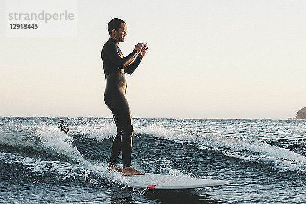 Seitenansicht in voller Länge von Surfer im Neoprenanzug filmt mit tragbarer Kamera auf Longboard