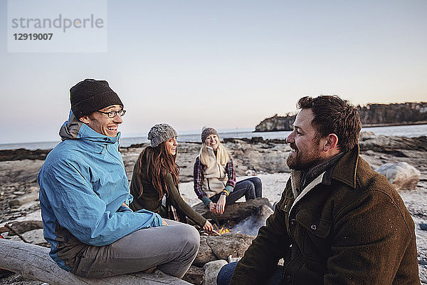 Vier erwachsene Freunde,  die sich am Lagerfeuer unterhalten und lächeln,  während sie an einem felsigen Küstenstrand im Herbst campen,  Portland,  Maine,  USA