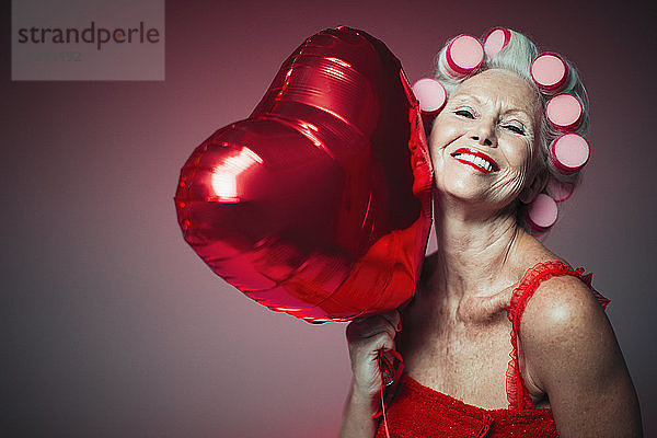 Porträt einer verspielten älteren Frau mit Lockenwicklern,  die einen Luftballon in Herzform hält