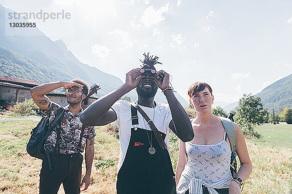 Drei junge Wanderfreunde schauen durch ein Fernglas aus der Feldlandschaft auf,  Primaluna,  Trentino-Südtirol,  Italien