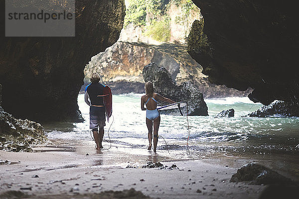 Rückansicht von Freunden,  die beim Strandspaziergang an Felsformationen ein Surfbrett tragen